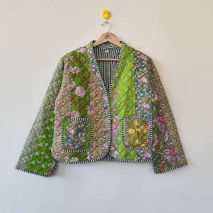 May include: A quilted jacket in shades of green with floral patterns and two front pockets. The jacket has a V-neck and is hanging on a wooden hanger. The lining is striped in black and white.