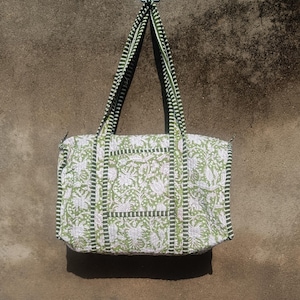 Pode incluir: Uma sacola com estampa floral verde e branca com alças listradas em preto e verde. A sacola é retangular com fecho de zíper e está pendurada em uma parede cinza texturizada.