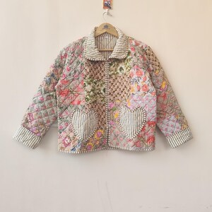 Könnte beinhalten: Gesteppte Jacke mit Patchwork-Design mit Blumenmustern und herzförmigen Taschen. Die Jacke hat einen gestreiften Kragen und Manschetten und hängt an einem Holzbügel. Die Farbpalette umfasst Rosa, Grün und Beige.