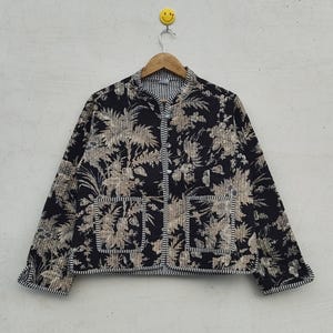 Op de afbeelding: Zwarte gewatteerde jas met een beige bloemenpatroon. De jas heeft een opstaande kraag, twee voorzakken en een gestreepte bies. Hij hangt aan een houten kleerhanger met een smiley-haak.