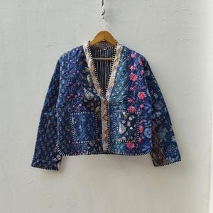 Pode incluir: Casaco acolchoado com design patchwork. Apresenta uma mistura de padrões florais e geométricos em tons de azul, rosa e branco. Tem fecho de botões, dois bolsos e está pendurado num cabide de madeira.