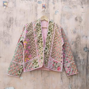 Puede incluir: Chaqueta acolchada con estampado floral en tonos rosa, verde y beige. La chaqueta tiene un forro a rayas rosas y ribetes rosas. Cuelga de una percha de madera contra una pared texturizada. El diseño floral es intrincado.