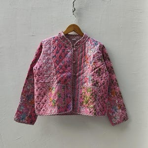 Könnte beinhalten: Gesteppte rosa Jacke mit floralem Muster. Die Jacke hat einen Stehkragen, lange Ärmel und eine Knopfleiste. Sie hängt an einem Holzbügel vor einer weißen Wand.