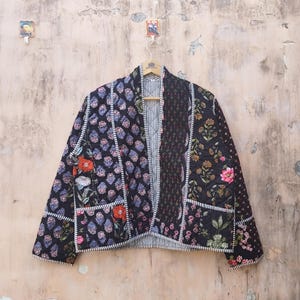 May include: A patchwork quilted jacket with a black background and floral and geometric patterns in red, pink, and blue. The jacket is hanging on a wooden hanger. The jacket has a striped lining.