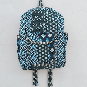 May include: A quilted backpack with a dark teal background and a repeating pattern of white and blue geometric shapes. The backpack has two side pockets, adjustable straps, and a top handle. The backpack is hanging on a white wall.