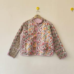 May include: A floral-patterned quilted jacket with a round collar and two front pockets. The jacket features a mix of pink, yellow, and green flowers on a light background. The edges are trimmed with pink piping.