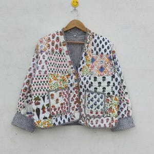 以下が含まれることがあります： 様々な色の花柄と幾何学模様のパッチワークデザインのキルティングジャケット。 ジャケットはVネック、ボタン留め、ストライプの袖口と裏地が付いています。 木製ハンガーに吊るされています。