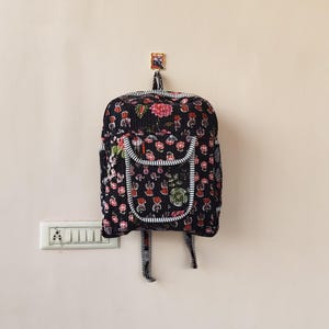 以下が含まれることがあります： 赤とピンクの花と緑の葉が特徴の、黒い花柄のバックパック。 バックパックにはフロントポケットと白と黒のストライプのトリムが付いています。 明るい色の壁に掛かっています。