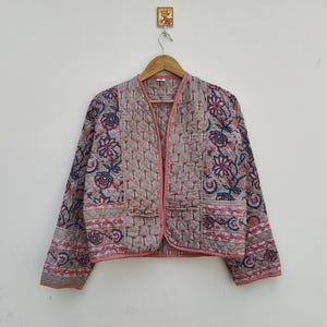 Peut inclure: Veste matelassée avec un motif floral dans les tons de rose, bleu et beige. La veste a un col en V et des manches longues. La veste a une bordure rose et deux poches.