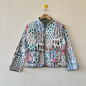 May include: A floral patterned quilted jacket in white, pink, blue, and green. The jacket has a mandarin collar, long sleeves, and a button-up front. It is hanging on a wooden hanger. The jacket's design features a variety of floral motifs.