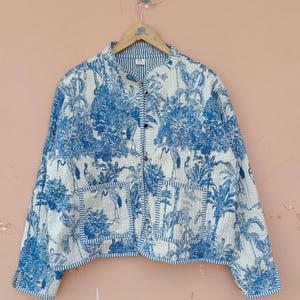 May include: A quilted jacket with a blue and white toile pattern featuring birds, trees, and other natural elements. The jacket has a mandarin collar, button closure, and two front pockets. The jacket is hanging on a wooden hanger.