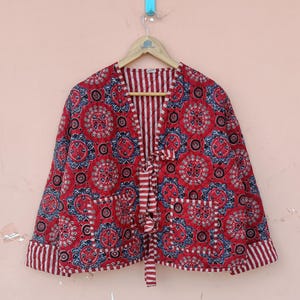 May include: A quilted jacket in a vibrant red hue, adorned with a detailed floral pattern in blue and white. The jacket features a striped interior and cuffs, with two front pockets and a tie closure. It hangs on a wooden hanger.