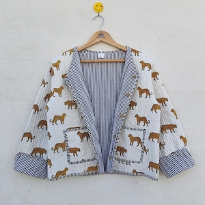 Könnte beinhalten: Eine weiße Steppjacke mit einem sich wiederholenden Muster aus braunen Tigern. Die Jacke hat ein marineblau-weiß gestreiftes Futter und Besatz. Die Jacke hängt an einem Holzkleiderbügel.