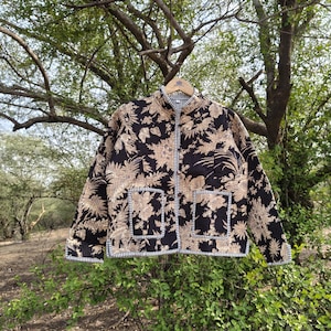 May include: A quilted jacket featuring a dark brown and beige floral pattern, a mandarin collar, long sleeves, and two front patch pockets. The garment is finished with a black and white striped trim along the edges, cuffs, and pocket openings.