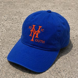 Puede incluir: Gorra de béisbol azul real con un diseño bordado en naranja. El diseño presenta las letras "NY" entrelazadas, con las palabras "SO" arriba y "OUTH" abajo, e "IC" a la derecha. La gorra tiene una visera curva y un botón superior.