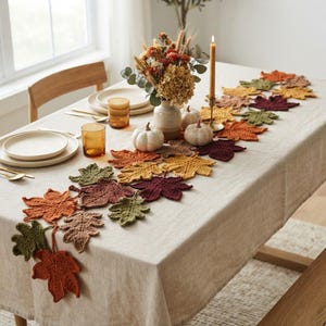 May include: A table runner composed of interconnected crocheted autumn leaves in shades of orange, red, brown, yellow, and green. The runner is laid across a light-colored tablecloth, accompanied by a vase of dried flowers and small pumpkins.