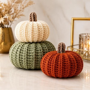 Puede incluir: Tres calabazas de ganchillo en color crema, verde salvia y naranja quemado. Cada calabaza presenta una textura acanalada y un pequeño tallo marrón texturizado en la parte superior. La calabaza crema está sobre la verde, con la naranja al lado.