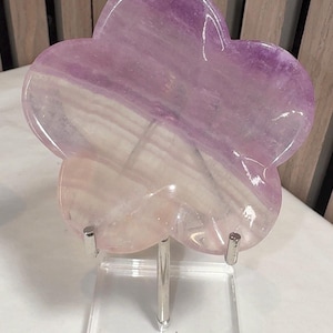 May include: A flower-shaped crystal sculpture in shades of purple and white, mounted on a clear acrylic stand with a silver metal post. The crystal has a polished, translucent appearance, showcasing natural patterns.