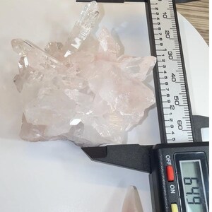 May include: A cluster of translucent, pale pink quartz crystals being measured by a black digital caliper. The display on the caliper clearly shows a measurement of 64.9 mm, indicating the width of the crystalline formation.