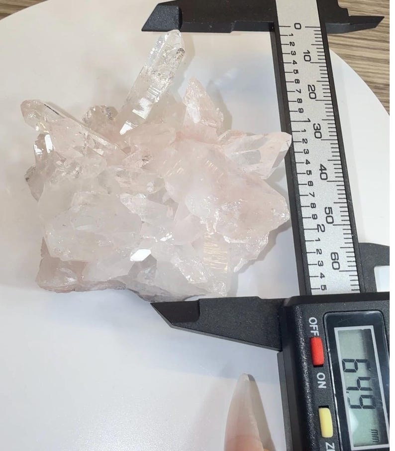 May include: A cluster of translucent, pale pink quartz crystals being measured by a black digital caliper. The display on the caliper clearly shows a measurement of 64.9 mm, indicating the width of the crystalline formation.