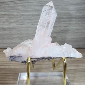 May include: A translucent, pale pink quartz crystal cluster featuring a prominent, elongated central point. The mineral specimen is mounted on a gold-toned metal wire stand, which is securely attached to a clear, rectangular acrylic base.