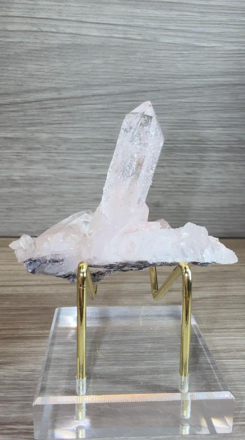 May include: A translucent, pale pink quartz crystal cluster featuring a prominent, elongated central point. The mineral specimen is mounted on a gold-toned metal wire stand, which is securely attached to a clear, rectangular acrylic base.