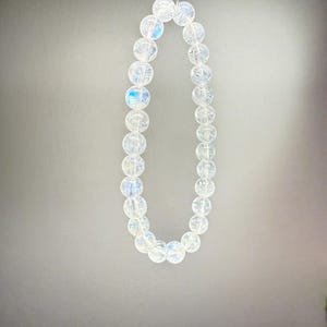 May include: A strand of clear, iridescent gemstone beads. The beads are round and strung together, creating a necklace or bracelet. The beads have a subtle, shimmering quality, reflecting light with hints of blue and other colors.