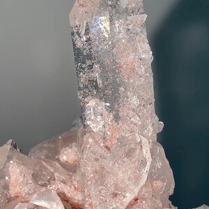 May include: A clear quartz crystal cluster featuring a prominent central point with translucent facets and internal inclusions. The base and surrounding smaller crystals exhibit a soft pink hue, with textured surfaces and sharp, angular edges throughout.