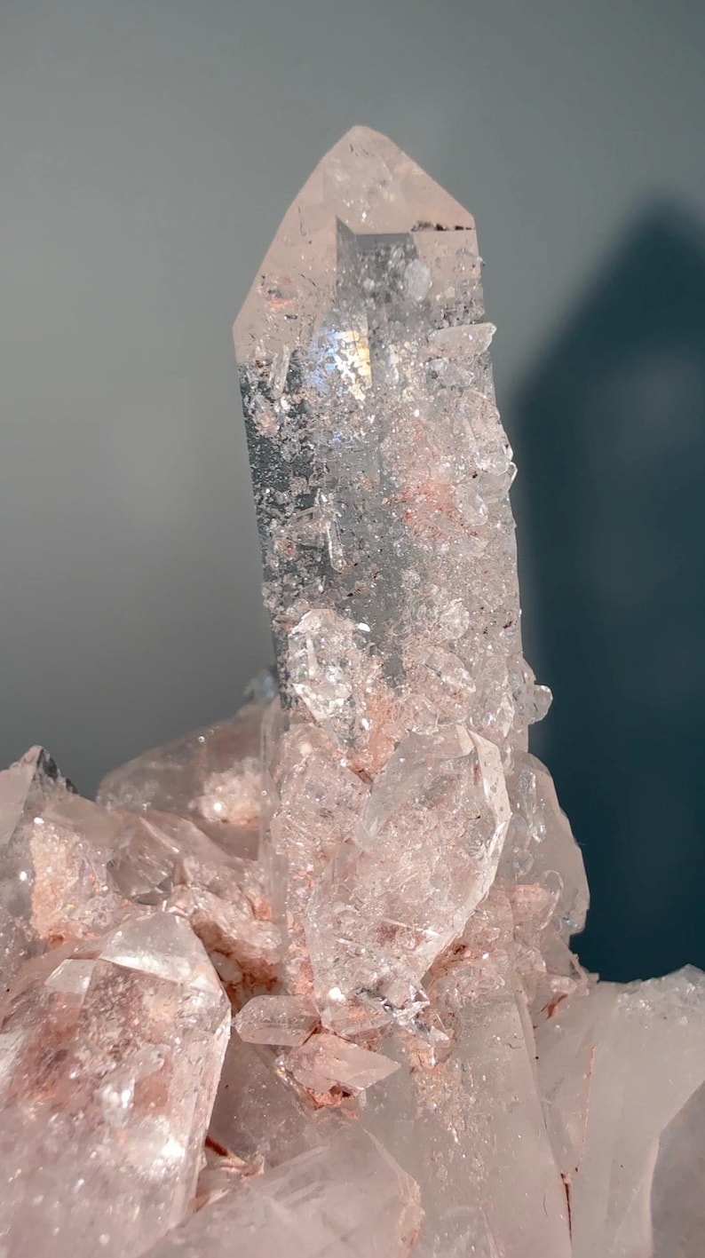 May include: A clear quartz crystal cluster featuring a prominent central point with translucent facets and internal inclusions. The base and surrounding smaller crystals exhibit a soft pink hue, with textured surfaces and sharp, angular edges throughout.