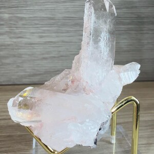 May include: A clear, pointed quartz crystal cluster with subtle pinkish inclusions, resting on a gold-toned metal wire stand attached to a transparent, square acrylic base, showcasing natural crystalline facets and sharp, translucent edges.