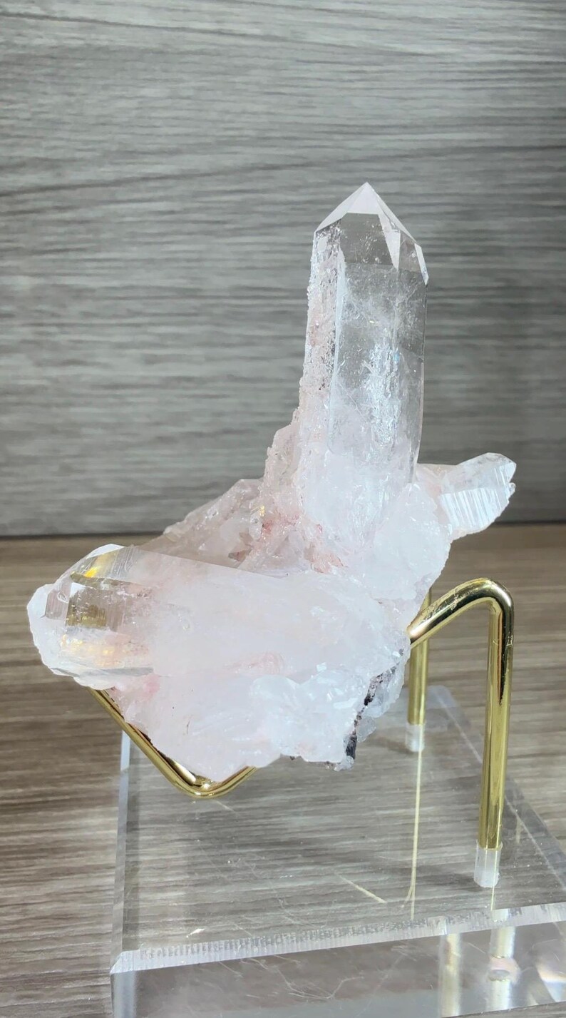 May include: A clear, pointed quartz crystal cluster with subtle pinkish inclusions, resting on a gold-toned metal wire stand attached to a transparent, square acrylic base, showcasing natural crystalline facets and sharp, translucent edges.
