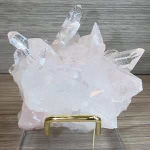 May include: A cluster of pale pink and translucent white quartz crystals with sharp, pointed terminations. The mineral specimen is supported by a gold-toned metal wire stand mounted on a clear, rectangular acrylic base for display purposes.
