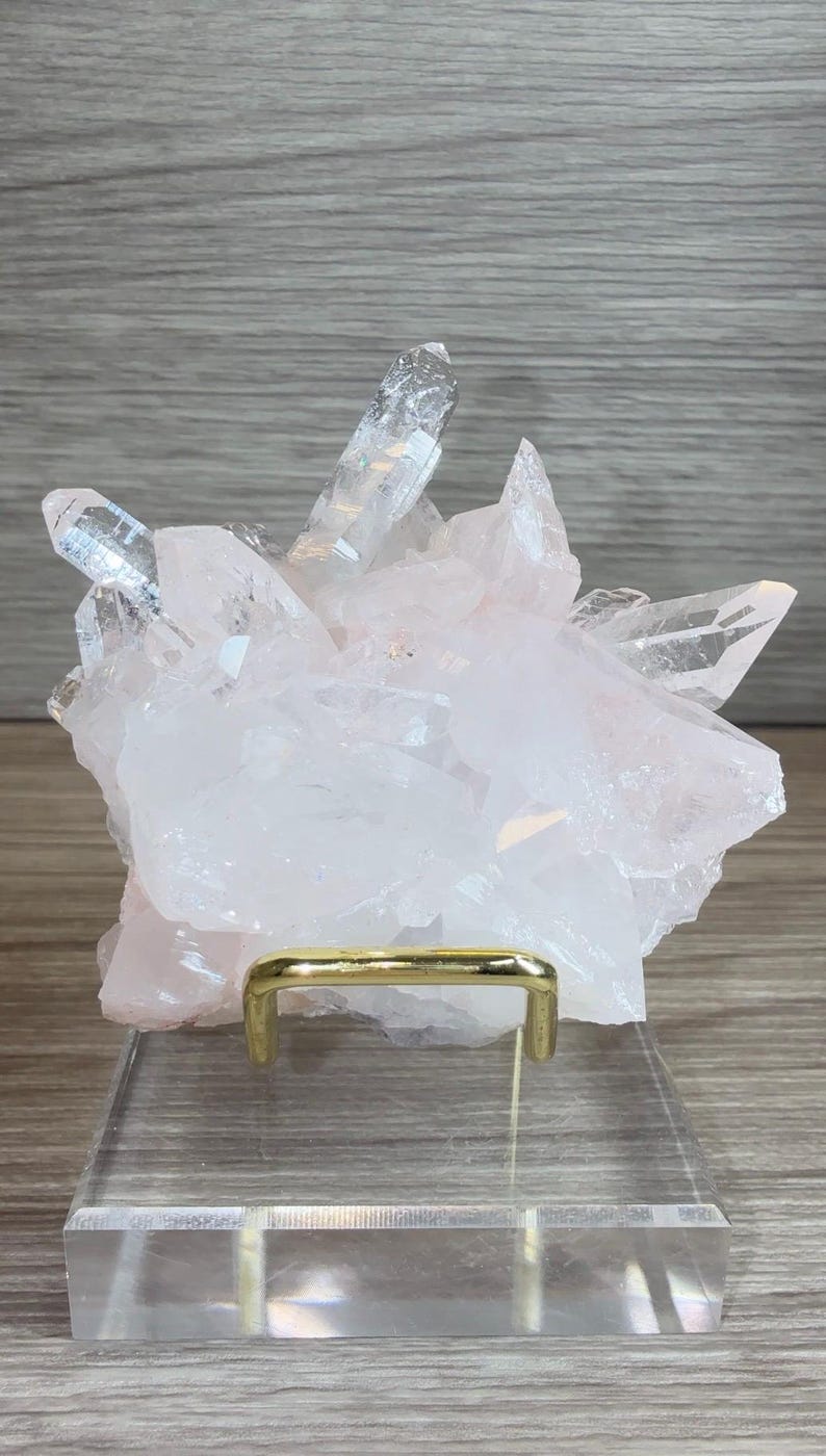May include: A cluster of pale pink and translucent white quartz crystals with sharp, pointed terminations. The mineral specimen is supported by a gold-toned metal wire stand mounted on a clear, rectangular acrylic base for display purposes.