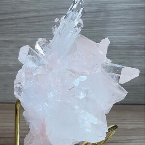 May include: A cluster of translucent, pale pink rose quartz crystals with sharp, pointed terminations. The mineral specimen is supported by a gold-toned metal wire stand attached to a clear, rectangular acrylic base for display purposes.