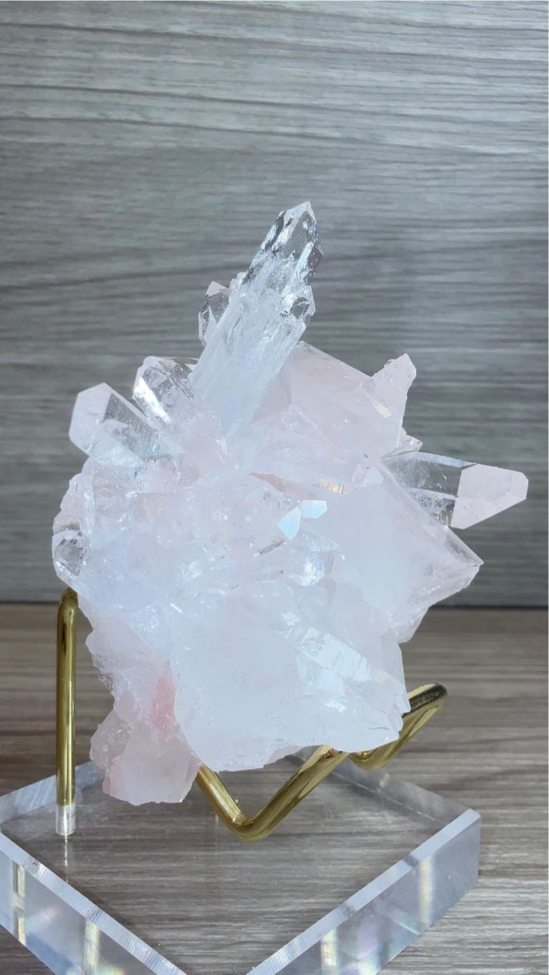 May include: A cluster of translucent, pale pink rose quartz crystals with sharp, pointed terminations. The mineral specimen is supported by a gold-toned metal wire stand attached to a clear, rectangular acrylic base for display purposes.