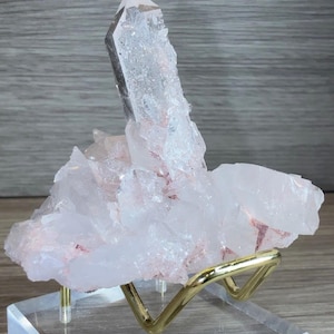 May include: A cluster of translucent, pale pink quartz crystals featuring a prominent, elongated central point. The mineral specimen is supported by a gold-toned metal stand mounted on a square, clear acrylic base, showcasing natural crystalline textures.