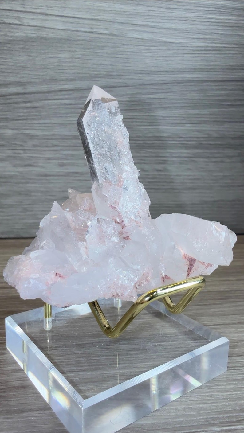 May include: A cluster of translucent, pale pink quartz crystals featuring a prominent, elongated central point. The mineral specimen is supported by a gold-toned metal stand mounted on a square, clear acrylic base, showcasing natural crystalline textures.