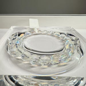 May include: A clear, iridescent beaded bracelet is displayed on a clear, circular stand. The bracelet has round, translucent beads that shimmer with a rainbow effect. The display is set on a white surface.