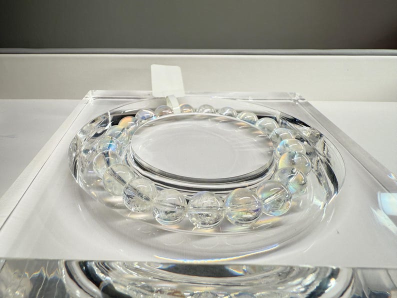 May include: A clear, iridescent beaded bracelet is displayed on a clear, circular stand. The bracelet has round, translucent beads that shimmer with a rainbow effect. The display is set on a white surface.