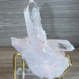 May include: A cluster of translucent, pale pink quartz crystals with a prominent vertical point, resting on a gold-toned metal stand attached to a clear acrylic base. The mineral specimen features jagged, crystalline edges and internal fractures.
