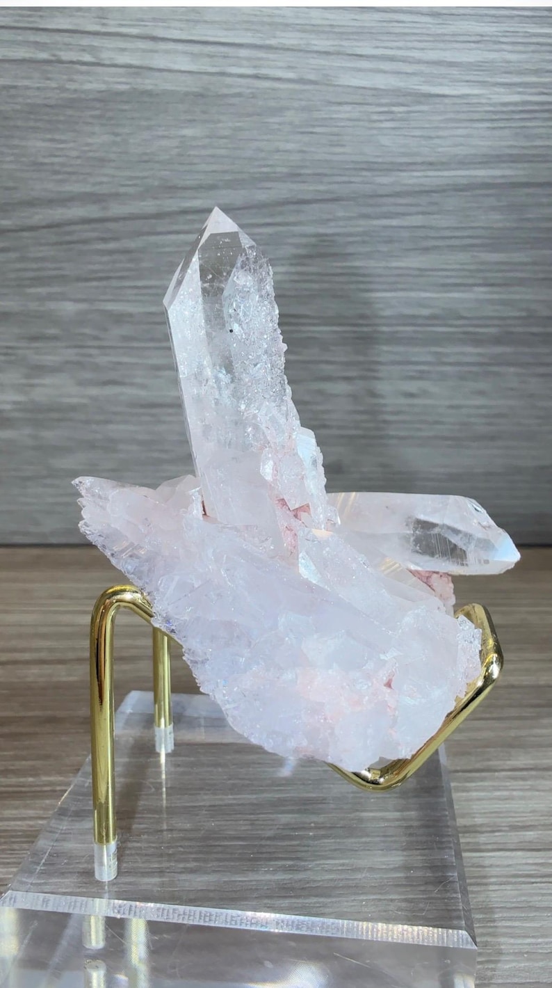 May include: A cluster of translucent, pale pink quartz crystals with a prominent vertical point, resting on a gold-toned metal stand attached to a clear acrylic base. The mineral specimen features jagged, crystalline edges and internal fractures.