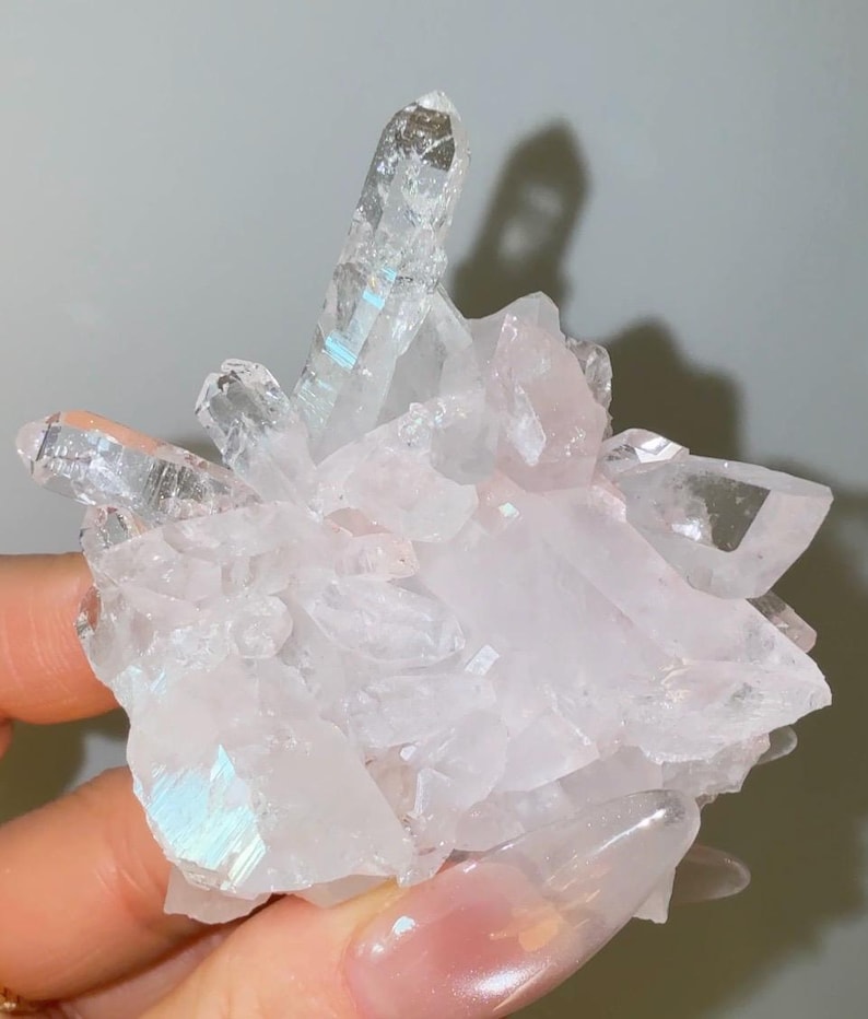 May include: A cluster of translucent, pale pink quartz crystals featuring a prominent, elongated central point. The mineral formation displays sharp, angular facets with subtle iridescent reflections and a clear, glassy texture throughout the structure.