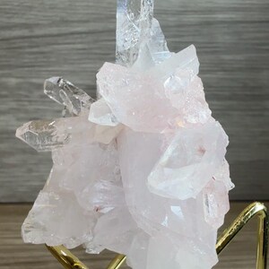 May include: A cluster of pale pink and translucent white quartz crystals with sharp, pointed terminations. The mineral specimen is supported by a gold-toned metal wire stand that features a clear acrylic base for stability and display.
