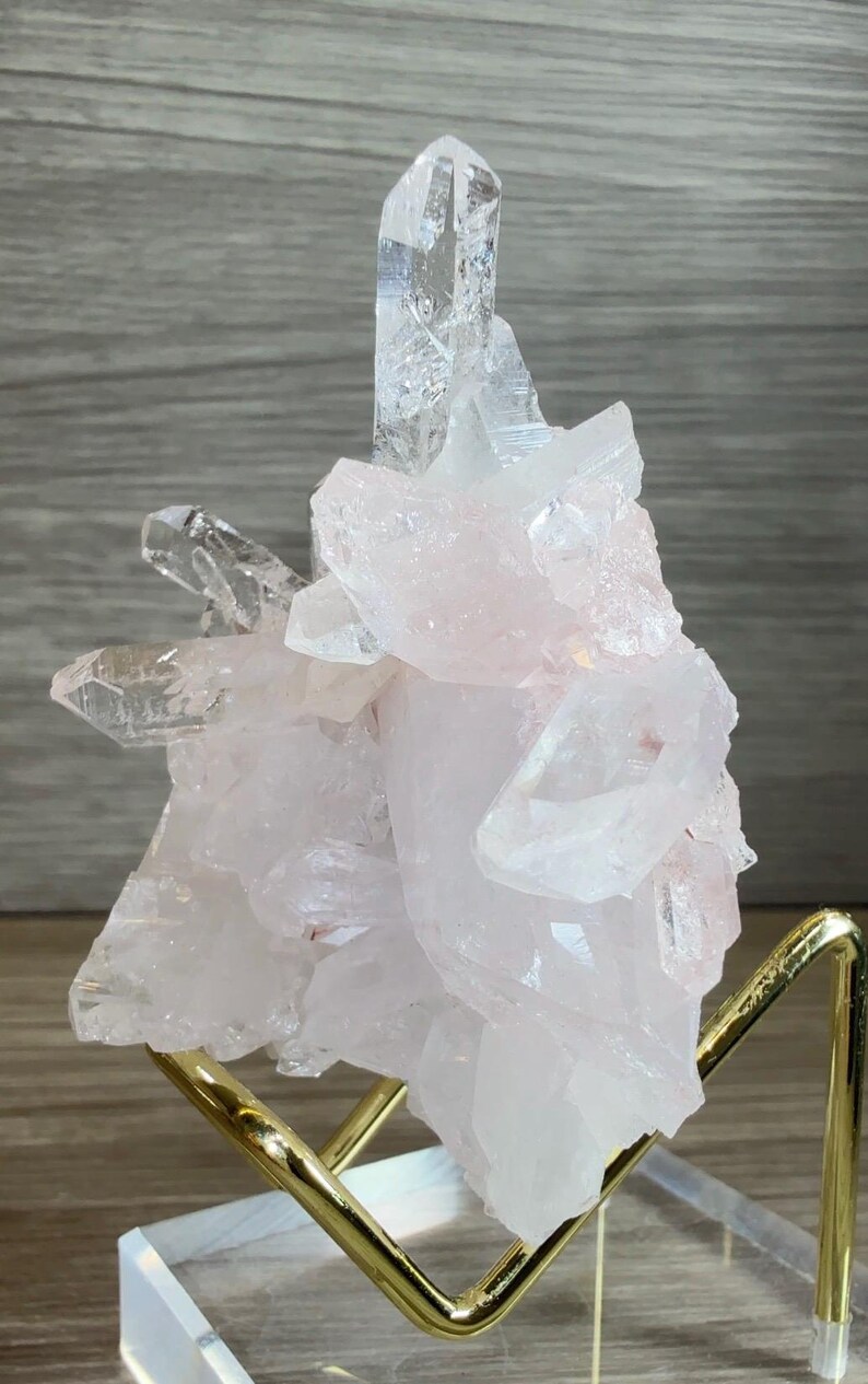 May include: A cluster of pale pink and translucent white quartz crystals with sharp, pointed terminations. The mineral specimen is supported by a gold-toned metal wire stand that features a clear acrylic base for stability and display.
