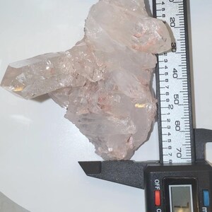 May include: A clear, jagged quartz crystal cluster with subtle pinkish inclusions. A black digital caliper is positioned against the crystal, showing a measurement of 71.0 mm on its display, indicating the width of the mineral specimen.