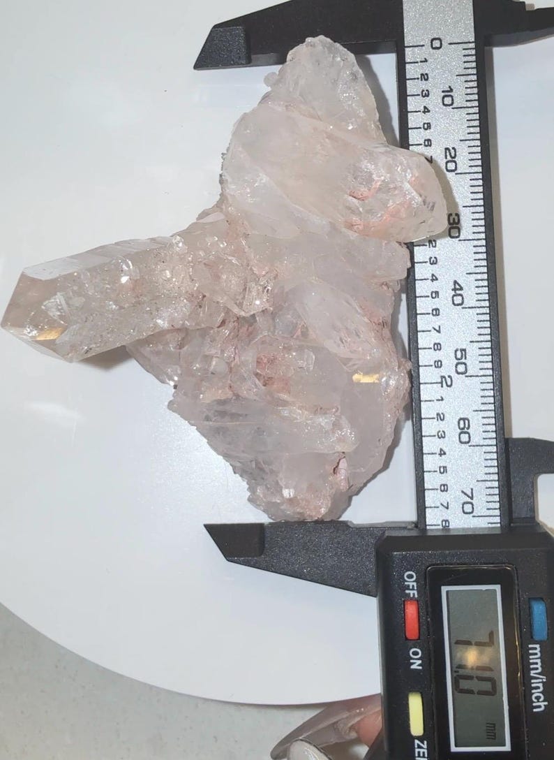May include: A clear, jagged quartz crystal cluster with subtle pinkish inclusions. A black digital caliper is positioned against the crystal, showing a measurement of 71.0 mm on its display, indicating the width of the mineral specimen.