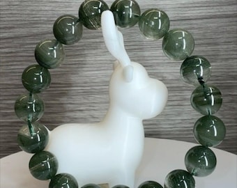 May include: A circular bracelet composed of translucent, dark green rutilated quartz beads, each measuring 10.5mm in diameter. The beads feature visible needle-like inclusions within the polished stone, strung together on an elastic cord.