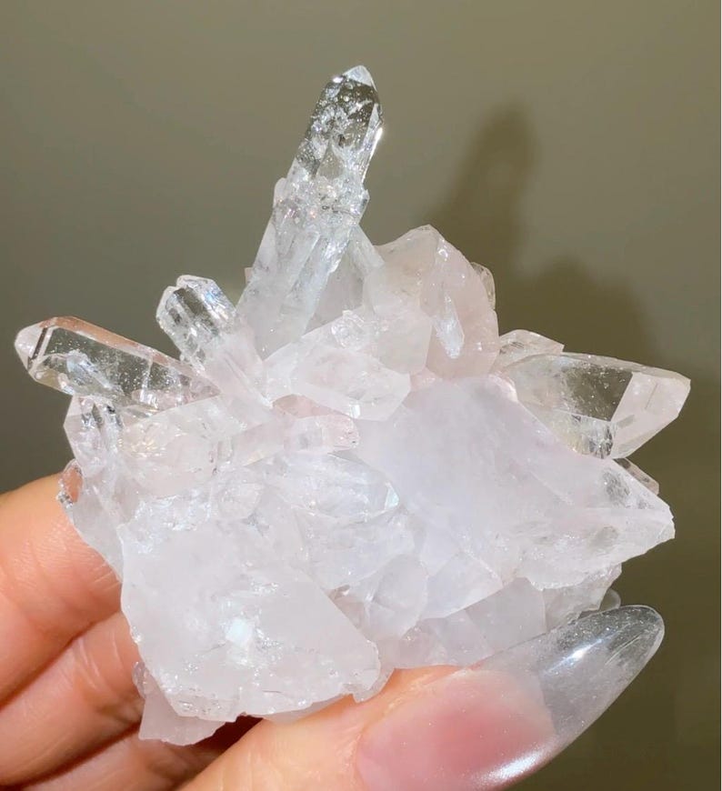 May include: A cluster of clear and pale pink quartz crystals featuring multiple sharp, pointed terminations extending from a central base. The mineral specimen displays a translucent, crystalline structure with varying facets and reflective surfaces.