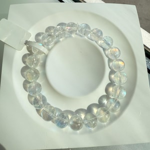 May include: A clear quartz bead bracelet with iridescent flashes. The bracelet is made of round, polished beads strung together. The beads are displayed on a white, square plate. The bracelet is a piece of jewellery.