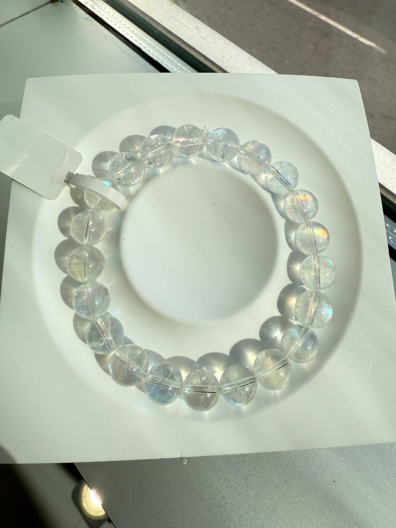 May include: A clear quartz bead bracelet with iridescent flashes. The bracelet is made of round, polished beads strung together. The beads are displayed on a white, square plate. The bracelet is a piece of jewellery.